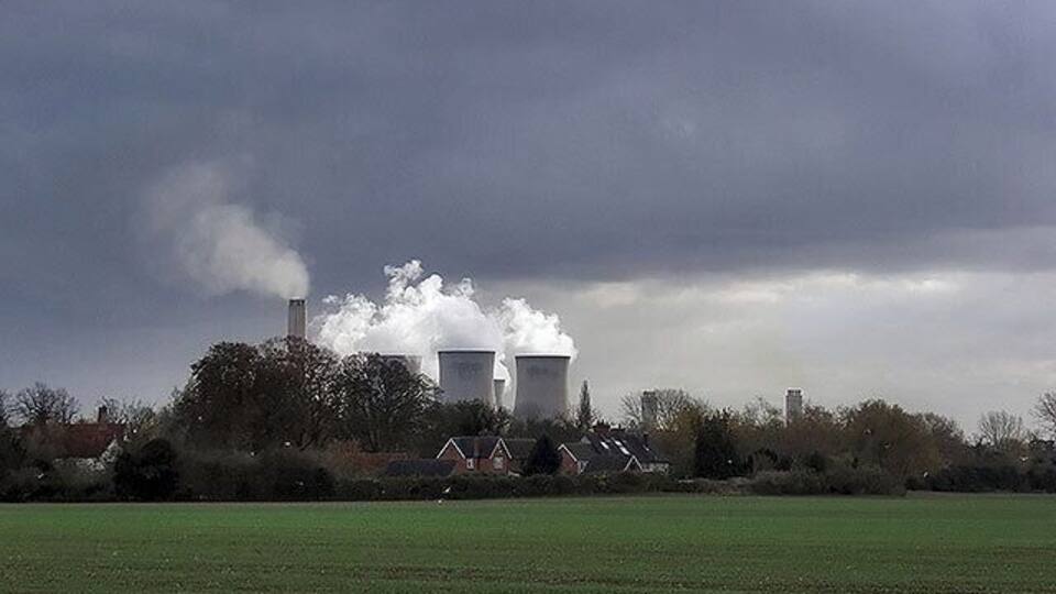A power plant with two cooling towers emitting steam, surrounded by fields and houses.