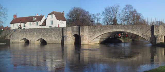 A stone bridge over a calm river, with a white building and trees in the background.