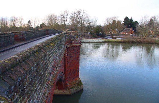 A stone bridge over a calm river, with a house and trees in the background.