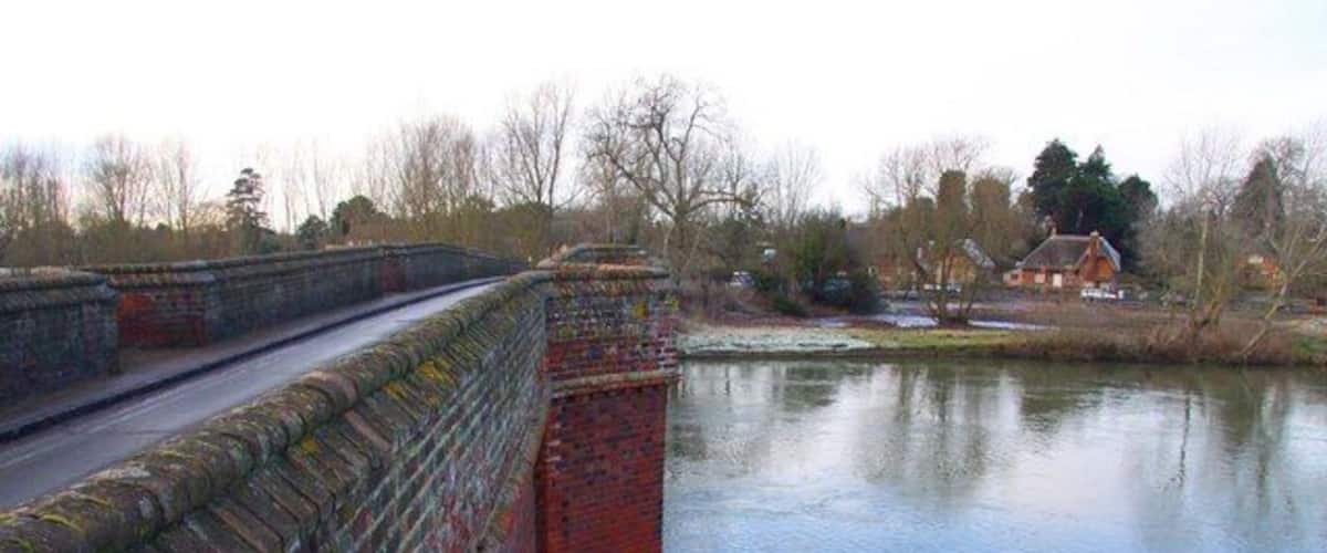 A stone bridge over a calm river, with a house and trees in the background.