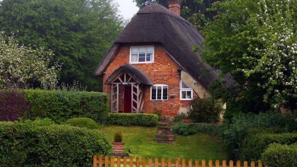 A thatched-roof cottage surrounded by lush greenery and a wooden fence.