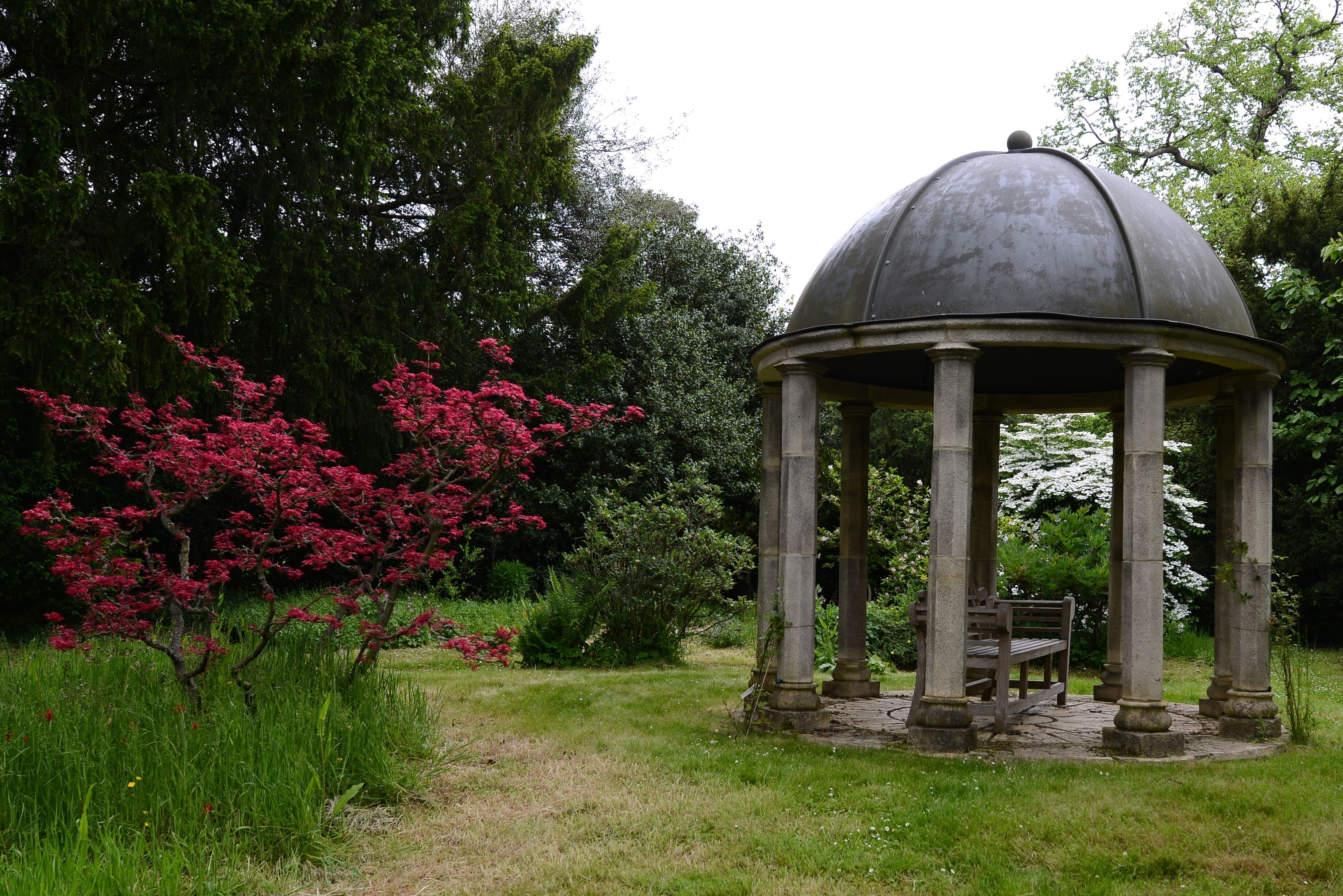A gazebo with a dome roof, surrounded by lush greenery and flowering trees.