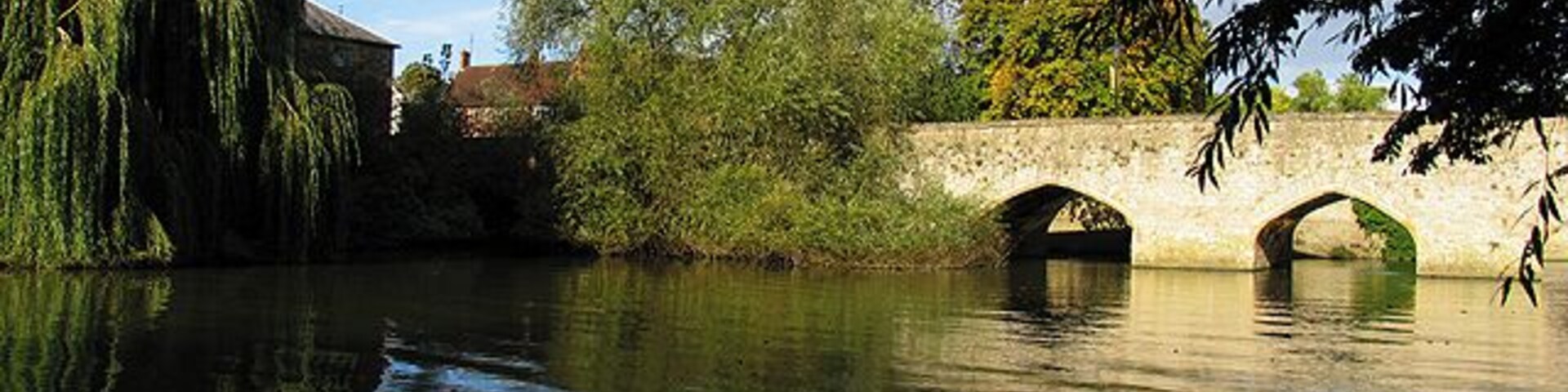 Un río tranquilo con un puente de piedra, árboles y un edificio al fondo.