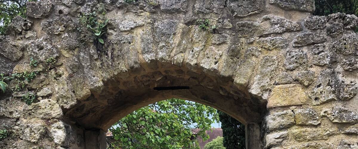 A stone archway with a metal gate, leading to a garden with flowers and a building in the background.