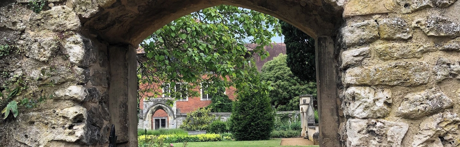 A stone archway with a metal gate, leading to a garden with flowers and a building in the background.