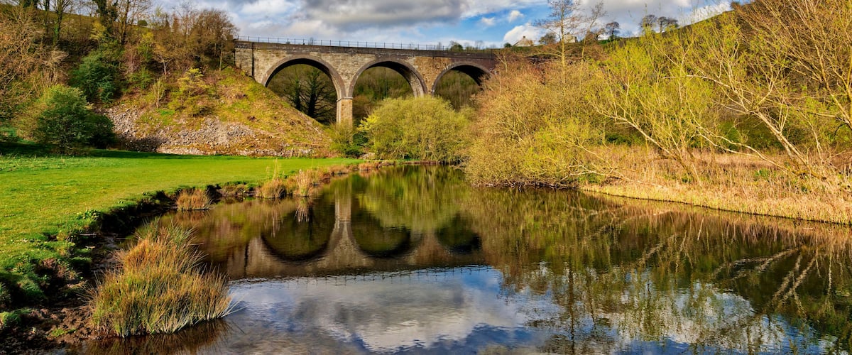 A stone bridge over a calm river with reflections, surrounded by trees and grassy banks.