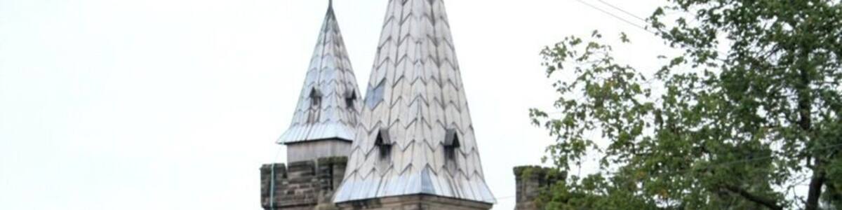 Ein historisches Steinhaus mit spitzem Dach und zwei Türmen, umgeben von gepflegten Hecken und einem Kiesweg.