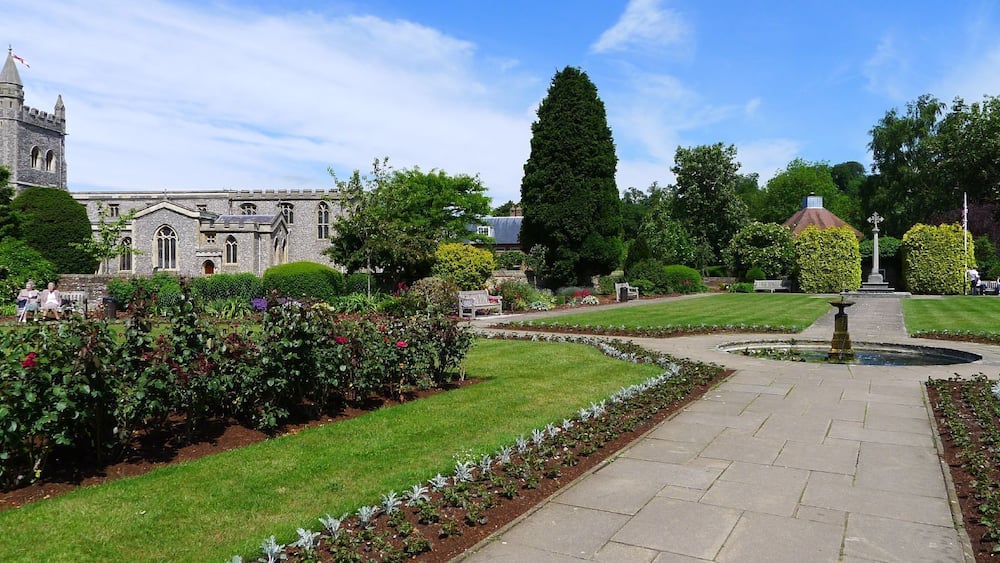 A historic building with a tower, a well-manicured garden with a fountain, and a clear blue sky.