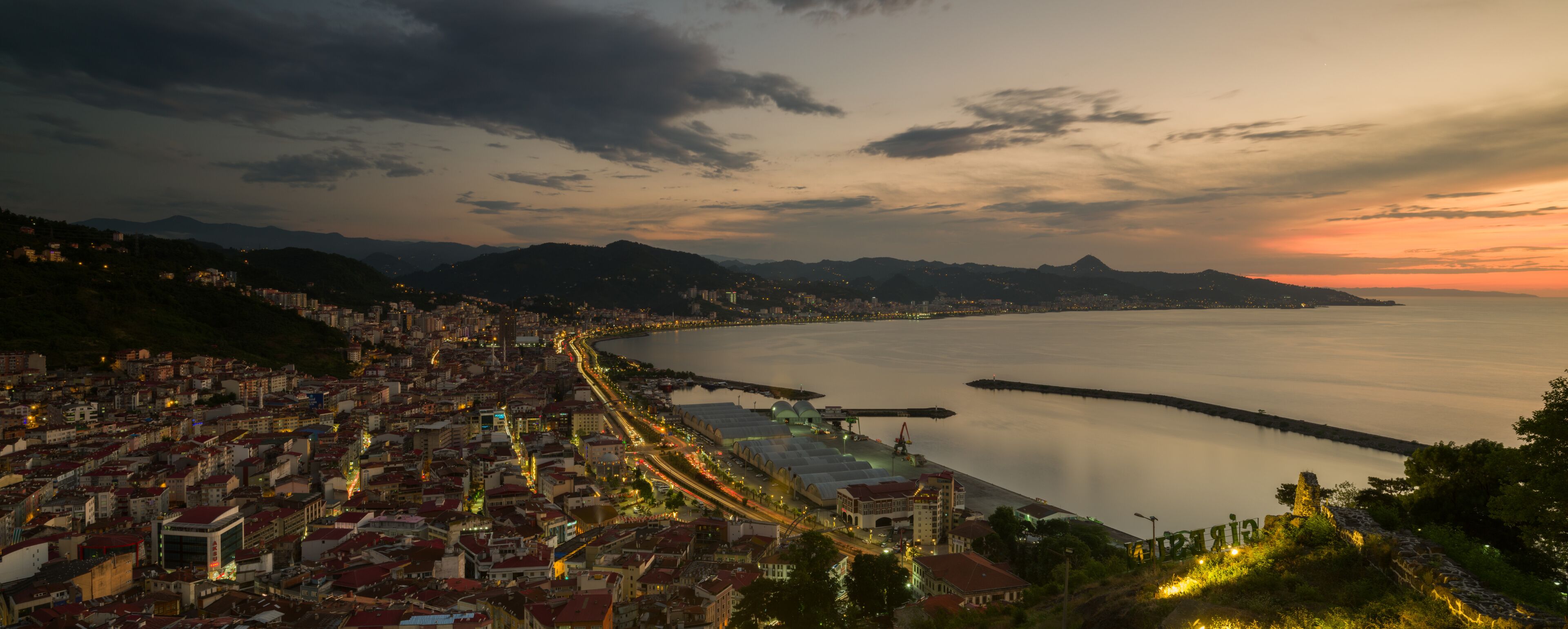 A coastal cityscape at dusk with a calm sea, illuminated streets, and distant mountains.