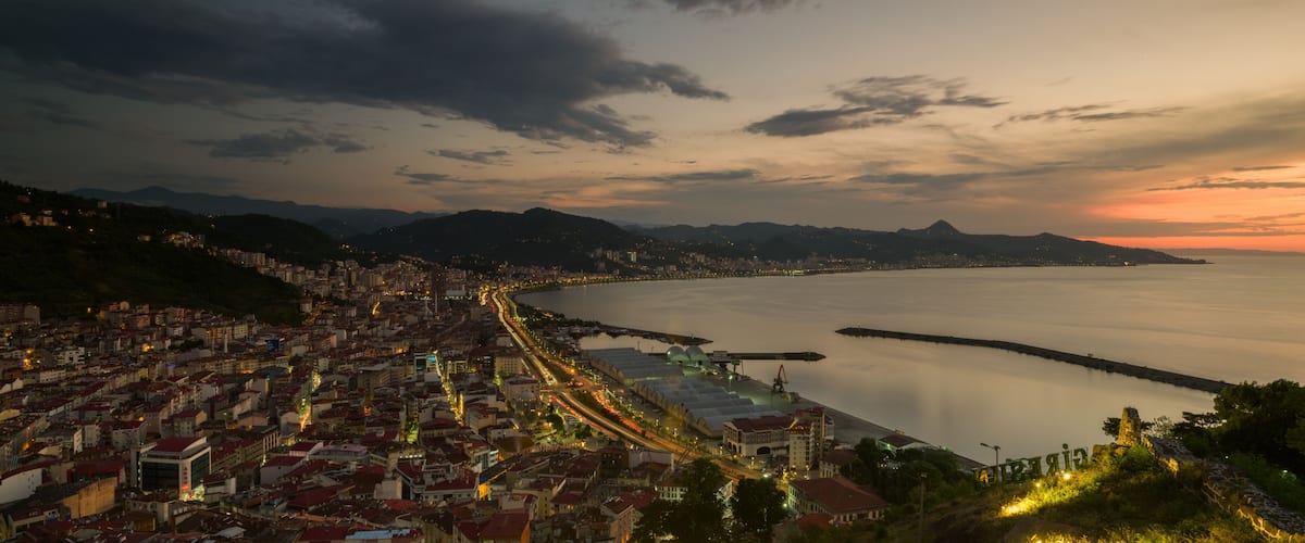 A coastal cityscape at dusk with a calm sea, illuminated streets, and distant mountains.