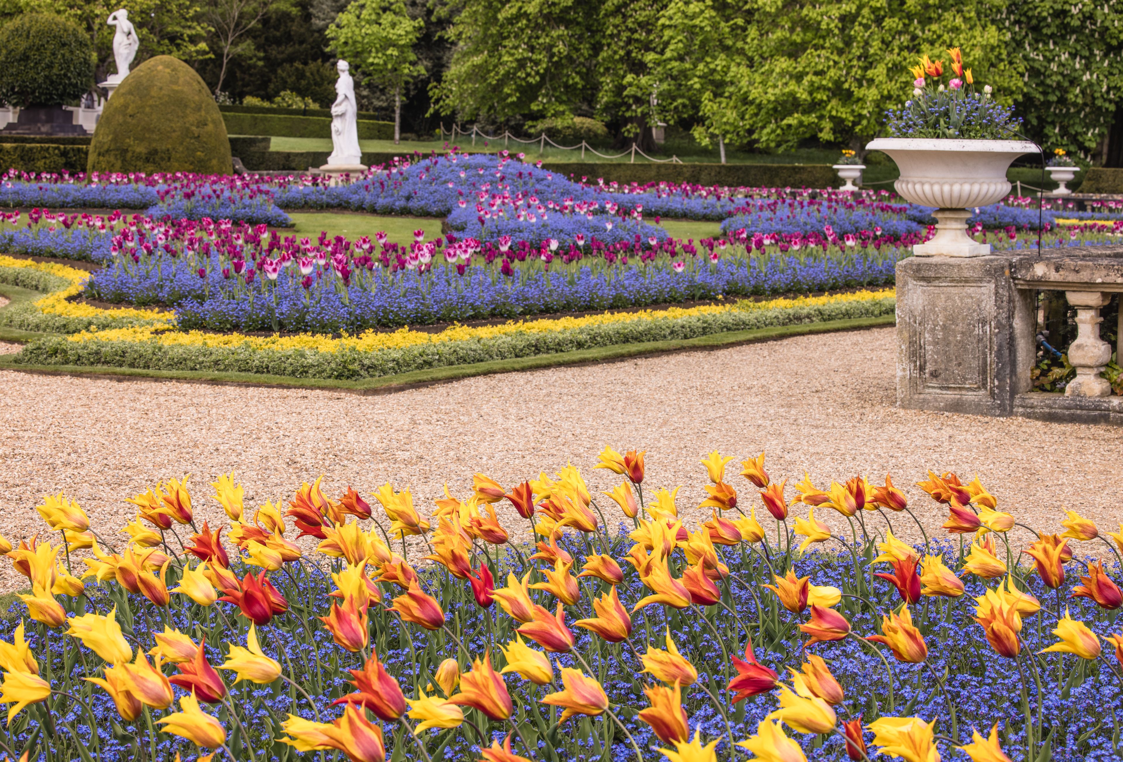 Een formele tuin met diverse kleurrijke bloemen, beelden en een fontein.
