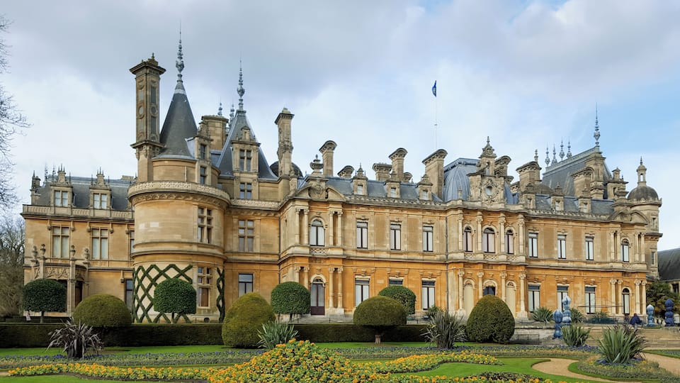 A grand historic building with ornate architecture, including multiple towers and a well-manicured garden with vibrant flowers.