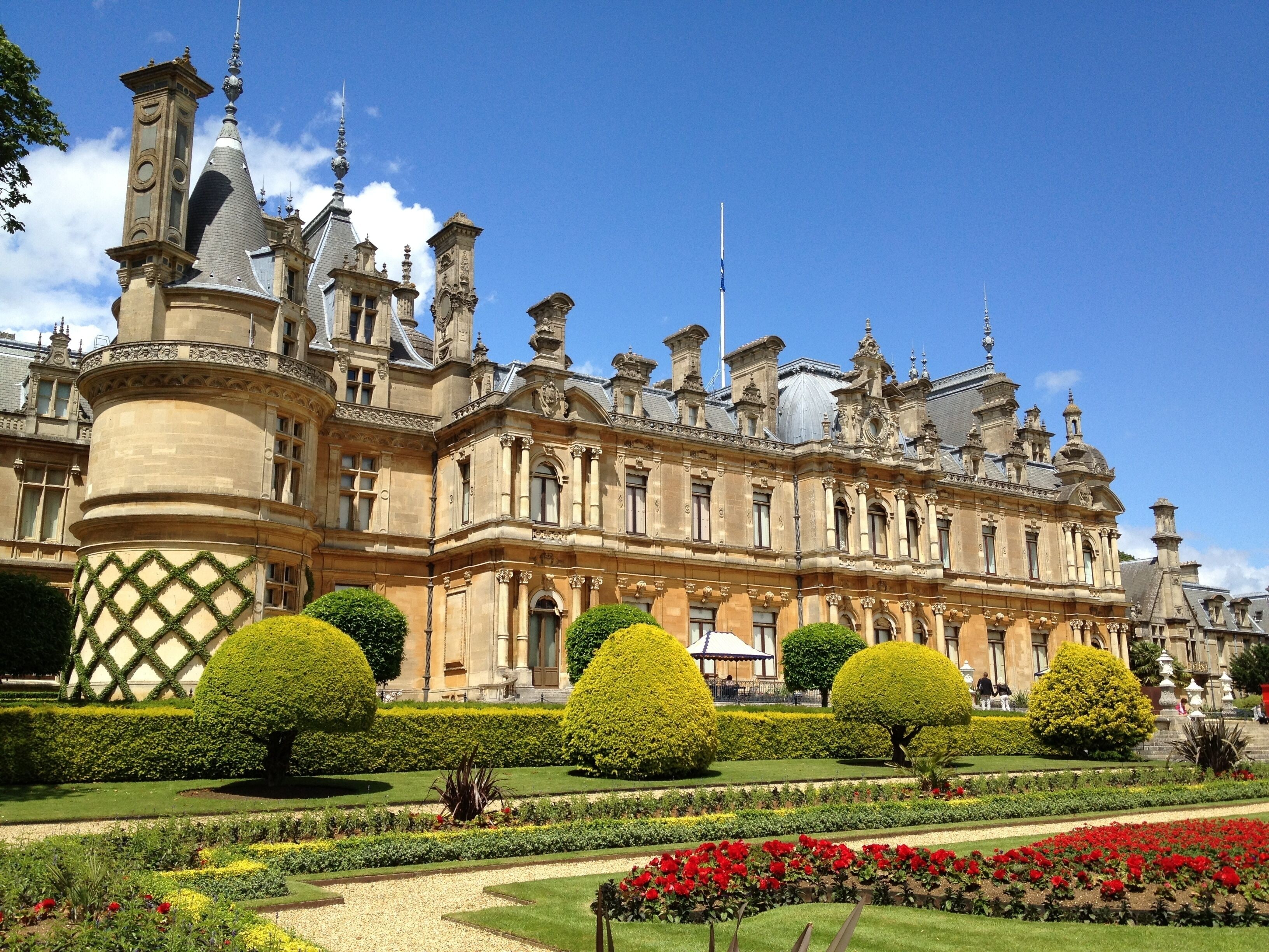 A grand historic building with ornate architecture, manicured gardens, and a clear blue sky.