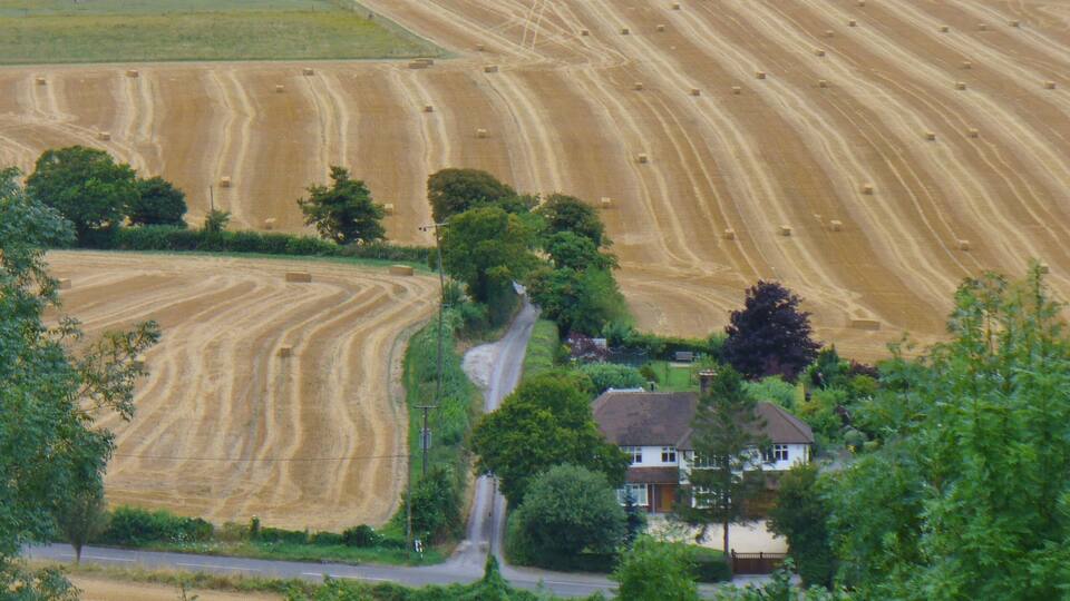 A rural landscape with fields of hay bales, a winding road, and a house surrounded by trees.