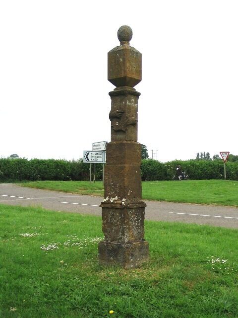Un monumento de piedra alto y desgastado con una parte superior esférica, situado junto a una carretera con señales de tráfico al fondo.