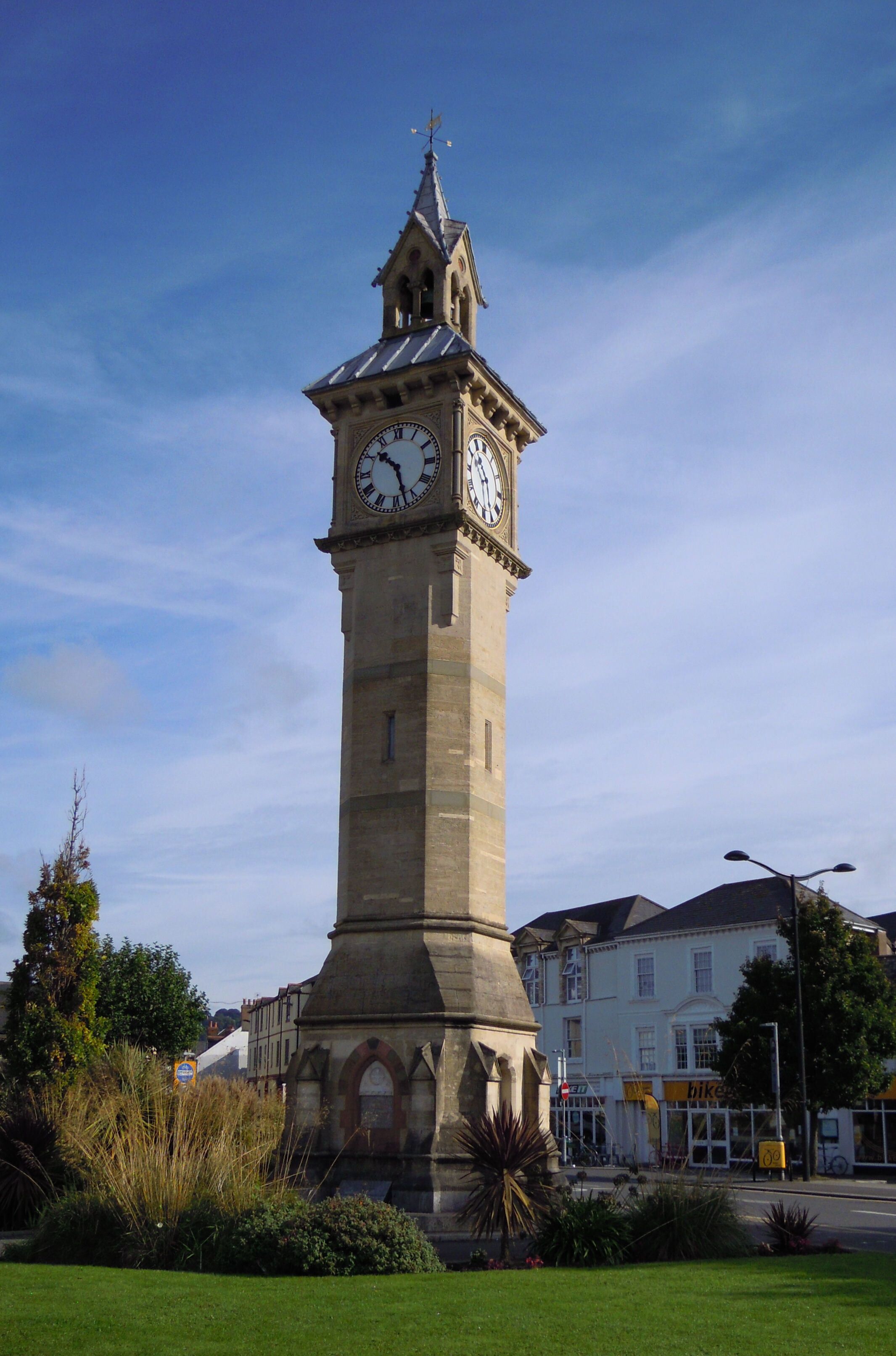 A tall clock tower with a pointed roof and a weather vane on top, surrounded by greenery and buildings in the background.