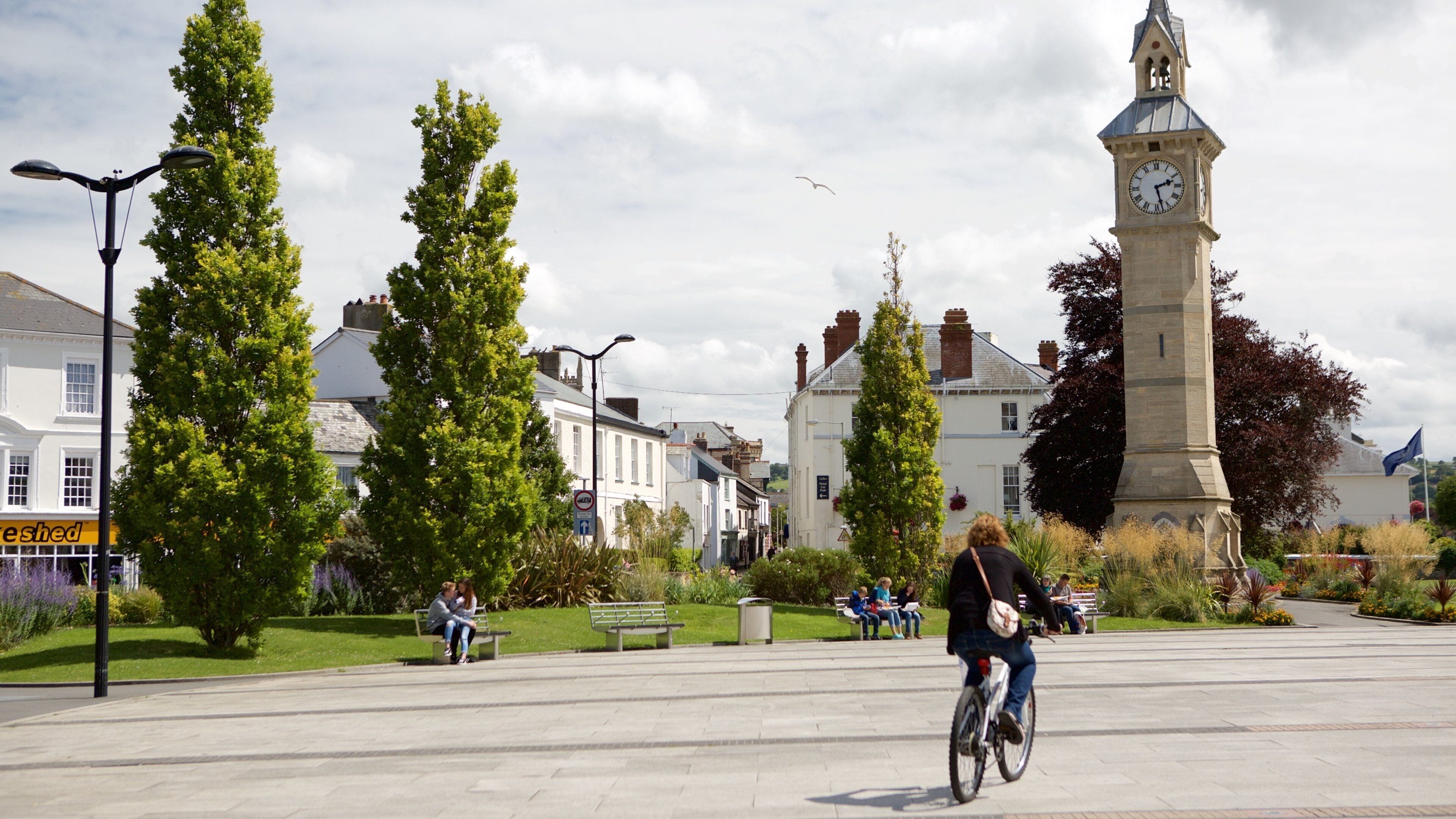 A town square with a clock tower, benches, cyclists, and greenery.
