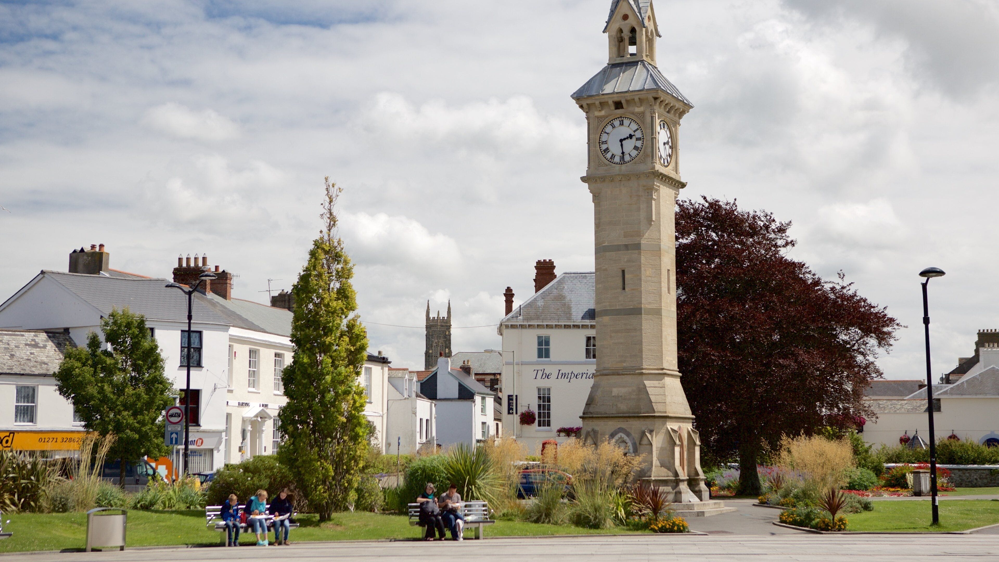 A clock tower in a town square with surrounding buildings and greenery.