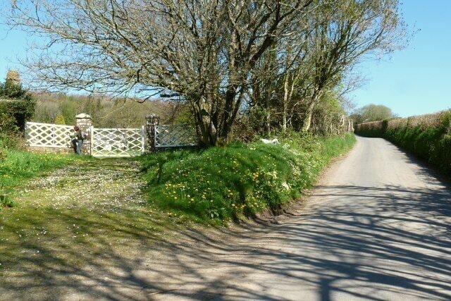 A narrow rural road flanked by trees and a gated entrance.