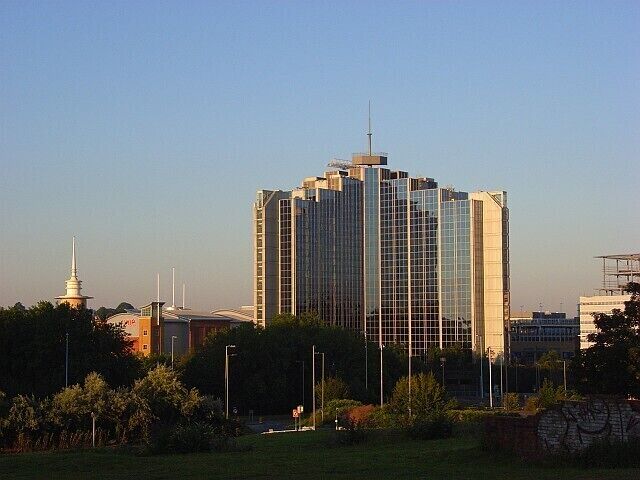 A modern high-rise building with a glass facade, surrounded by greenery and a few smaller structures in the background.