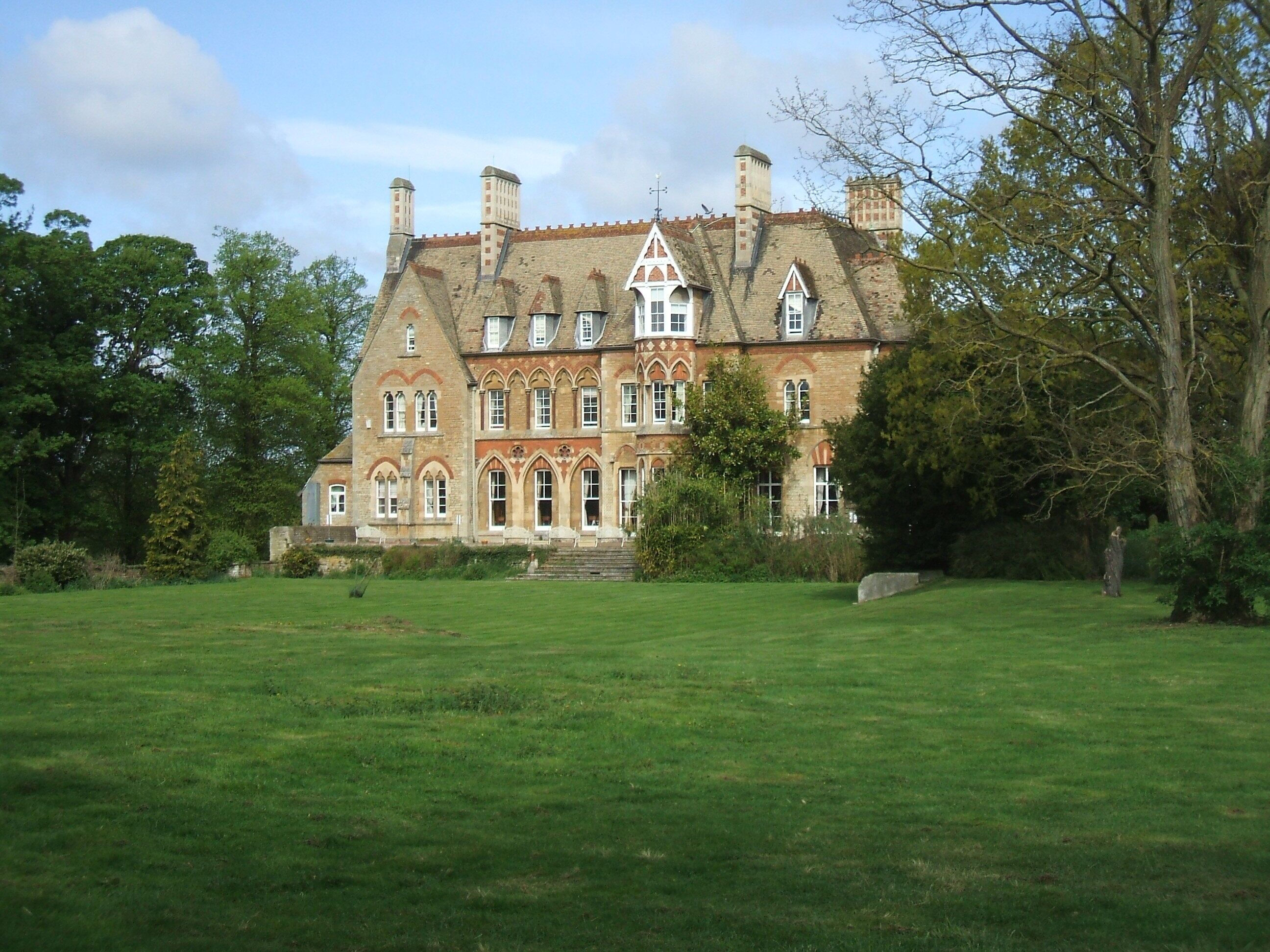 A large, historic building with a prominent tower and multiple chimneys, surrounded by lush green lawns and trees.