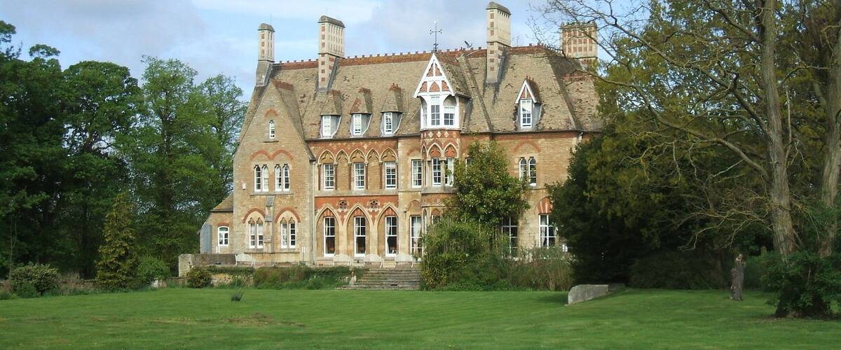 A large, historic building with a prominent tower and multiple chimneys, surrounded by lush green lawns and trees.