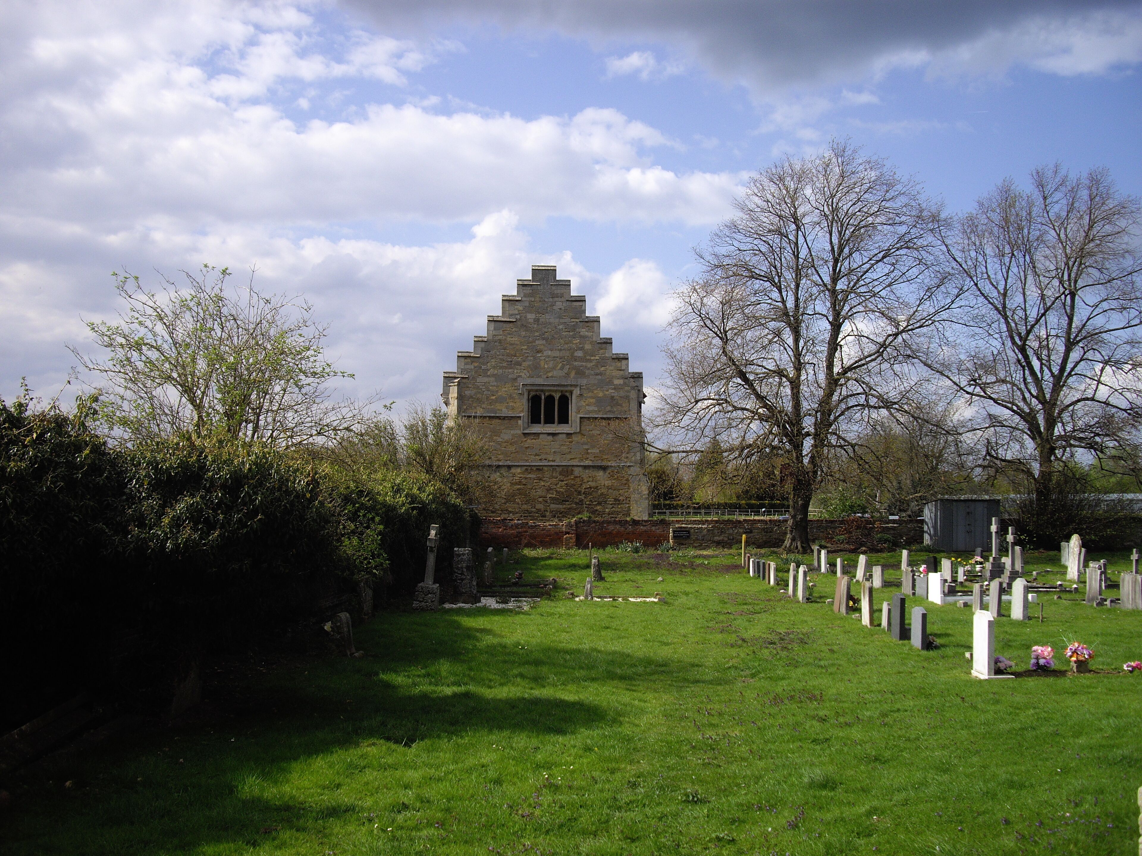 A stone church with a triangular roof and a small window, surrounded by a cemetery with headstones and a grassy path.