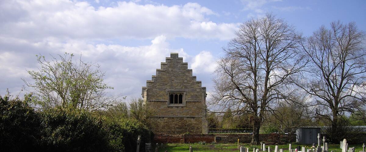 A stone church with a triangular roof and a small window, surrounded by a cemetery with headstones and a grassy path.