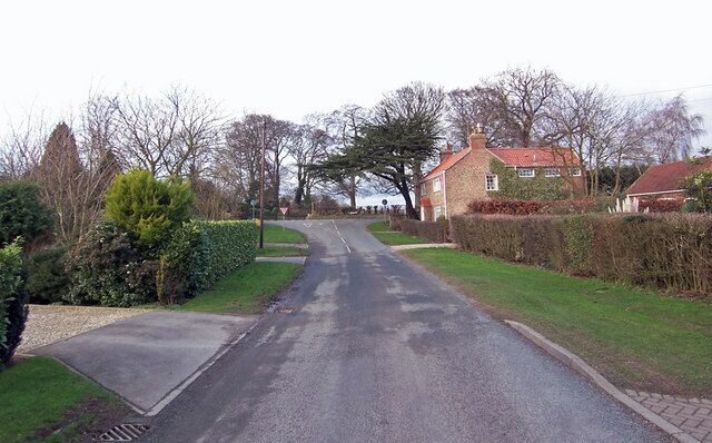 A quiet residential street with houses on both sides, surrounded by greenery and trees.
