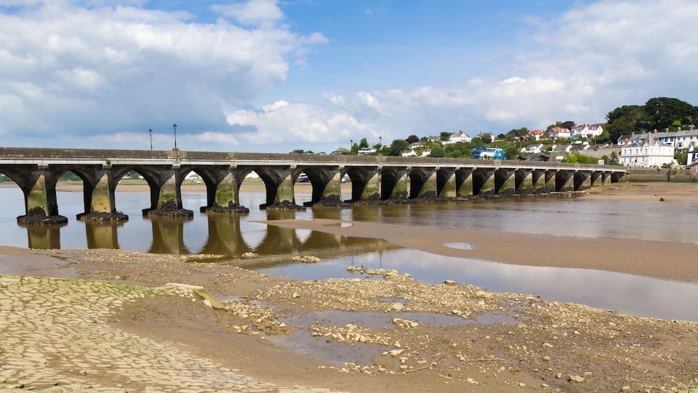 A stone bridge over a river with a sandy beach in the foreground and houses in the background.