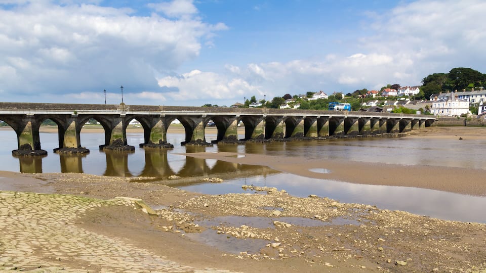 A stone bridge over a river with a sandy beach in the foreground and houses in the background.