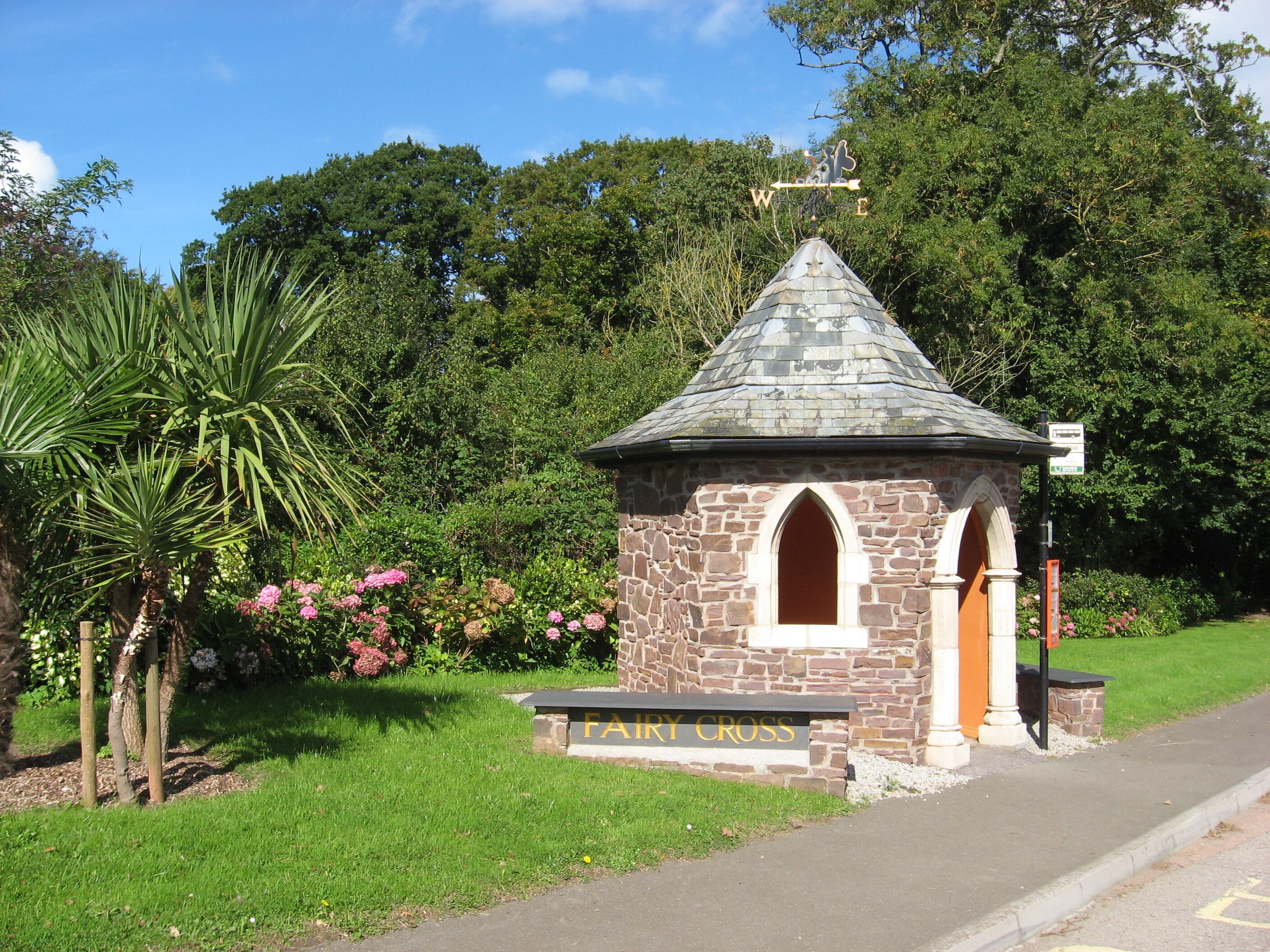 A stone structure with a pointed roof and a sign reading "Fairy Cross" in a lush garden.