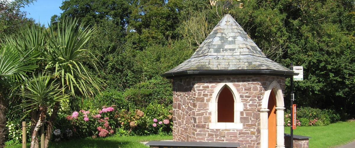 A stone structure with a pointed roof and a sign reading "Fairy Cross" in a lush garden.