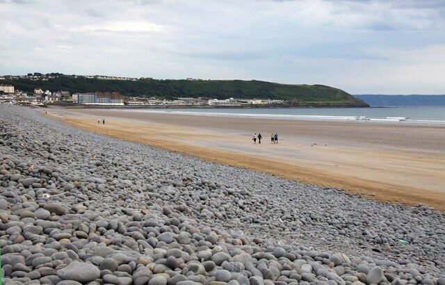 A pebbled beach with people walking, a distant hill, and buildings on the shore.
