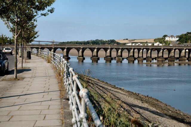 A stone bridge over a river, a paved walkway with a railing, and a few parked cars.