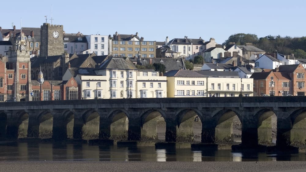 A stone bridge with arches spanning a river, with historic buildings and a clock tower in the background.