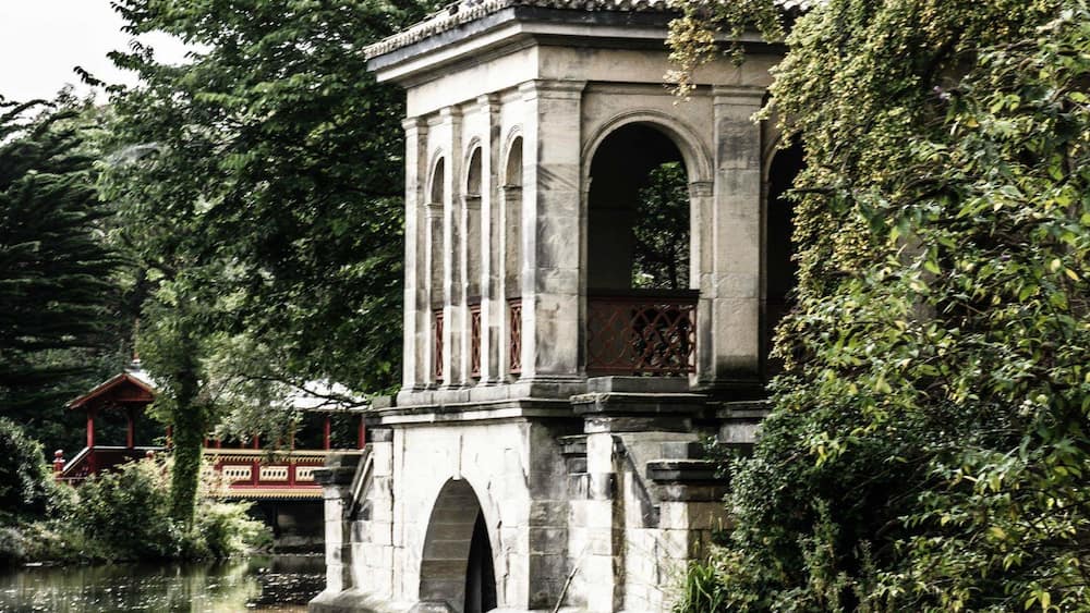 Ein steinerne Pavillon mit Bögen und einem Ziegeldach, umgeben von üppigem Grün und einem Gewässer.