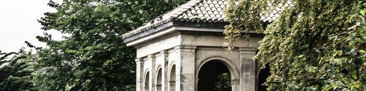 Ein steinerne Pavillon mit Bögen und einem Ziegeldach, umgeben von üppigem Grün und einem Gewässer.