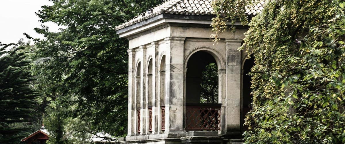 Un pavillon en pierre, doté d’arches et d’un toit en tuiles, entouré d’une végétation luxuriante et bordé par un plan d’eau.