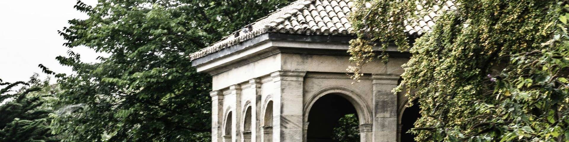 A stone pavilion with arches and a tiled roof, surrounded by lush greenery and a body of water.