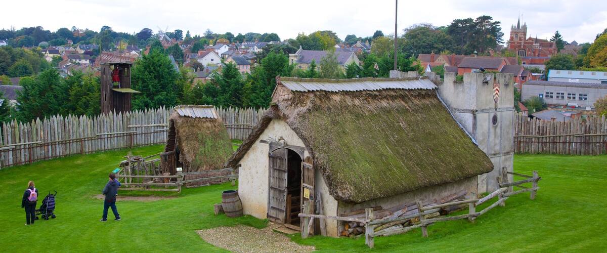 A historical village with thatched-roof buildings, a wooden fence, and a flagpole.