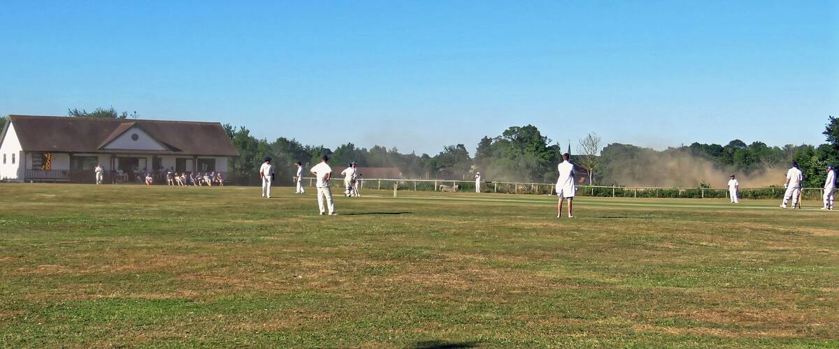 A cricket field with players, a scoreboard, and a building in the background.