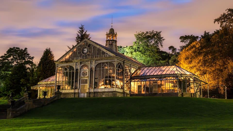 A large, ornate greenhouse with a clock tower, surrounded by lush greenery and trees.
