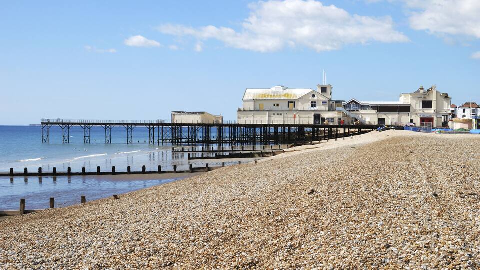A pebbled beach with a pier extending into the sea, and buildings along the shore.
