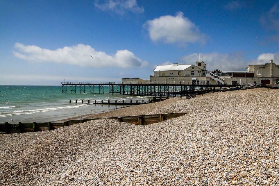 A pebble beach with a pier extending into the sea, a building with a sloped roof, and a clear sky.