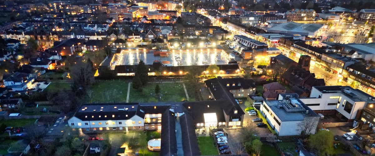 An aerial view of a residential area with houses, a parking lot, and a large building with illuminated windows.