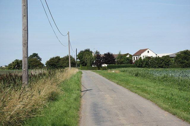 A rural road with a white house on the right, surrounded by fields and trees.