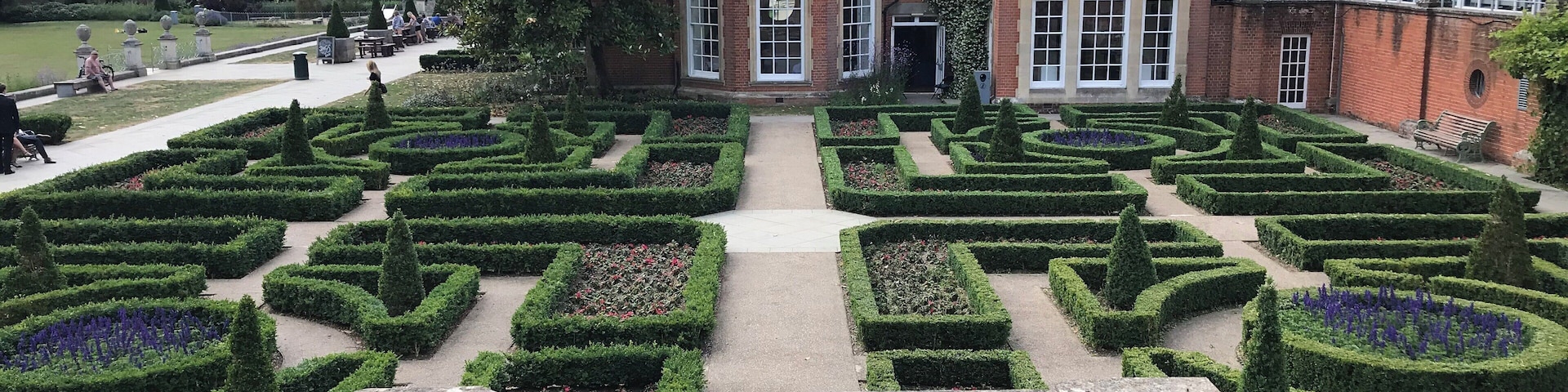 A formal garden with geometric hedges and a large brick building in the background.