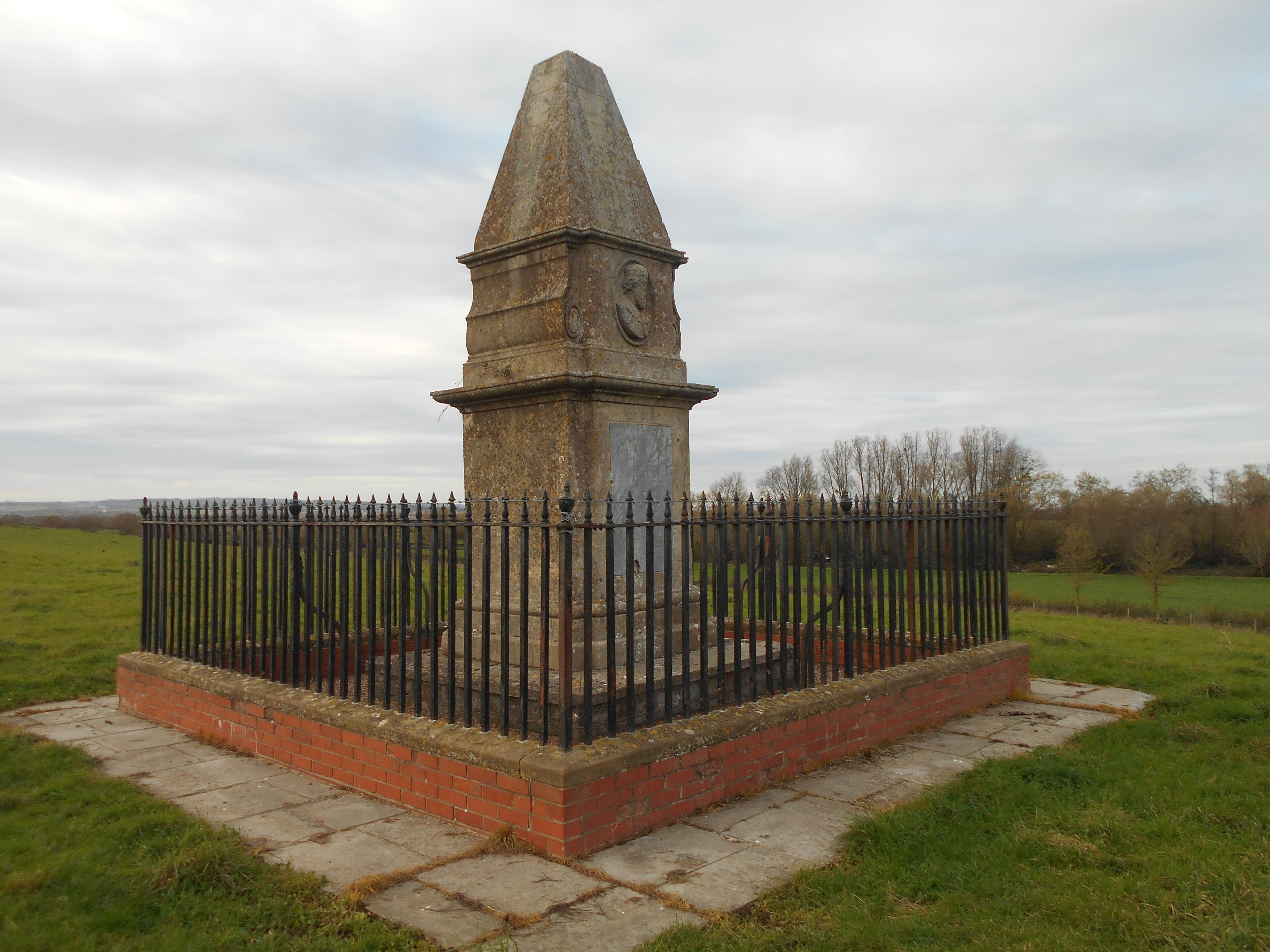 A weathered stone monument with a clock, surrounded by a brick base and a metal fence in a grassy field.