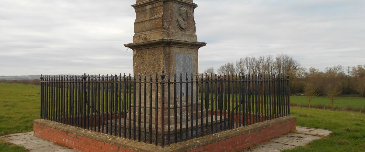 A weathered stone monument with a clock, surrounded by a brick base and a metal fence in a grassy field.
